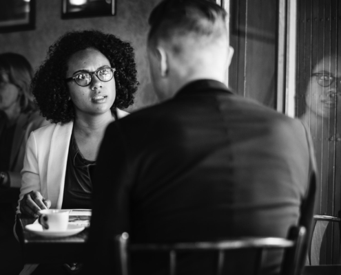 Woman sitting in a cafe facing a man with angry look whose back is to camera.