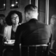 Woman sitting in a cafe facing a man with angry look whose back is to camera.