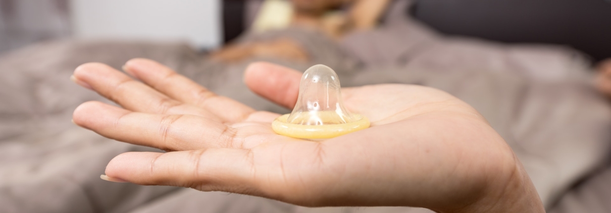 Condom sitting on top of a hand with background image showing a man under a bed sheet